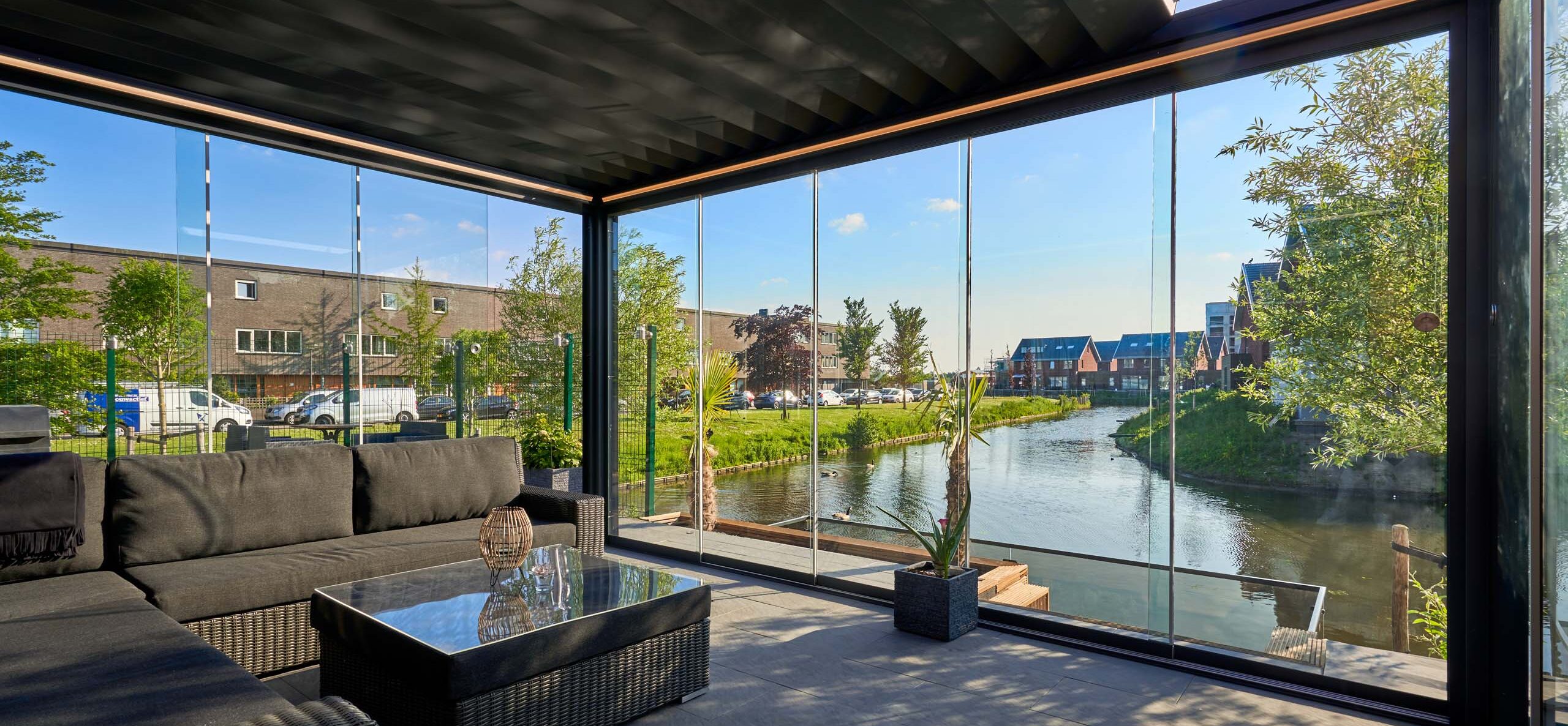 Moderne overkapping met glazen schuifwanden aan het water, ingericht met een donkere loungebank en tafel, met uitzicht op een rustige woonwijk en groen langs de oever.